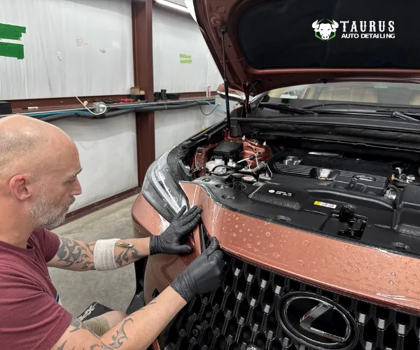 A technician wearing black gloves carefully applies a clear protective film to the front trim of a bronze luxury vehicle with the hood open.
