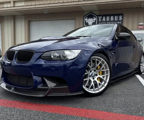Low-angle front side view of a dark blue BMW M3 coupe parked in front of Taurus Auto Detailing.